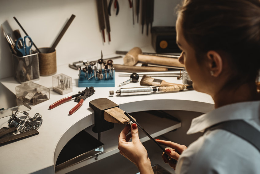 Focused on a process. Back view of a female jeweler working and shaping an unfinished ring with a tool at workbench in workshop.